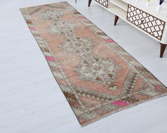 Puede incluir: Una alfombra rectangular de aspecto envejecido, con un motivo floral central en tonos melocotón, marrón y crema. La alfombra presenta un borde con diseños geométricos y toques de rosa brillante. Esta alfombra de estilo vintage es adecuada para un pasillo o entrada.