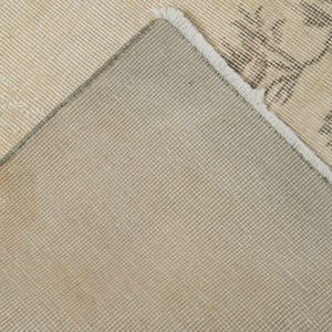 May include: Close-up of a beige and gray area rug with a floral pattern. The rug has a distressed look, with visible wear and tear. The underside of the rug is a solid beige color, and the edges are frayed.