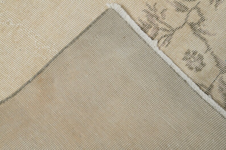 May include: Close-up of a beige and gray area rug with a floral pattern. The rug has a distressed look, with visible wear and tear. The underside of the rug is a solid beige color, and the edges are frayed.