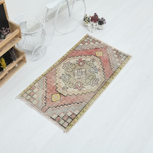 May include: A rectangular area rug featuring a central geometric medallion in shades of muted red, beige, and grey. The rug has a repeating diamond pattern border with small yellow accents and fringed edges on the short sides of the textile.