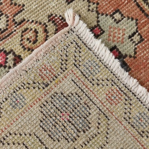 May include: Close-up of the woven edge of a rug with a fringe. The rug has a beige background with a multi-colored geometric pattern.
