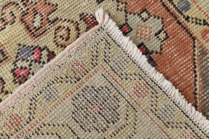 May include: Close-up of the woven edge of a rug with a fringe. The rug has a beige background with a multi-colored geometric pattern.