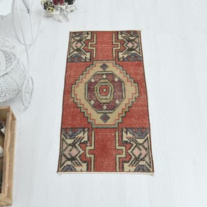 May include: A rectangular area rug featuring a central geometric medallion in shades of red, beige, and dark blue. The rug displays a symmetrical pattern with intricate borders and angular motifs, showcasing a weathered texture and a flat-woven design.