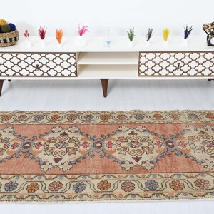 May include: A vintage runner rug with a peach background and intricate floral patterns in shades of beige, brown, and blue. The rug has a distressed look and is perfect for adding a touch of bohemian style to any room.