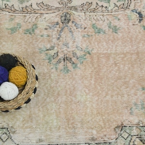 May include: A woven basket containing five small balls of yarn in black, blue, yellow, and white, placed on a large, faded, light-colored rug featuring a repeating floral pattern in muted green, brown, and black tones.