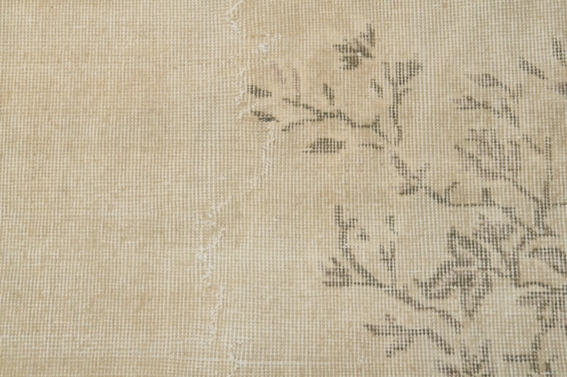 May include: Close-up of a beige rug with a floral pattern in shades of brown and gray. The rug has a textured surface and a distressed appearance, suggesting an antique or vintage style. The design is visible on the right side.