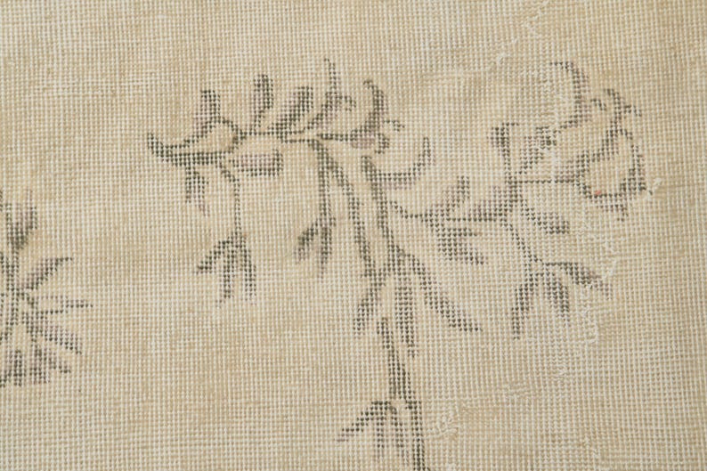 May include: Close-up of a beige rug with a floral pattern in shades of gray. The rug has a textured surface and a faded, vintage appearance. The floral design is spread across the rug.
