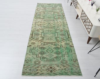 May include: A rectangular, green and brown patterned rug with a central geometric design. The rug has a distressed, vintage appearance and is placed on a white floor. The rug's design features repeating geometric shapes and borders.
