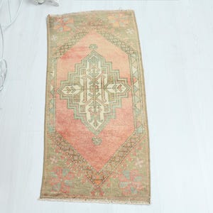 May include: A rectangular, vintage-style rug with a central diamond pattern in shades of peach, green, and beige. The rug has a distressed look, with a faded appearance and fringed edges. The rug is approximately 24 inches long and 12 inches wide.