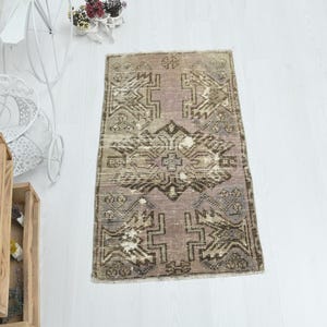 May include: A rectangular area rug featuring a muted mauve and brown color palette with a central geometric medallion and symmetrical angular motifs throughout the field. The rug displays a distressed texture with visible variations in pile density.