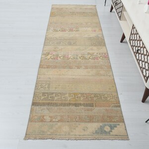May include: A rectangular, beige runner rug with a distressed, vintage appearance. The rug features horizontal stripes with faded floral and geometric patterns in muted tones of pink, green, and brown. The rug is approximately 10 feet long.
