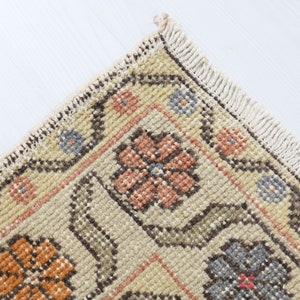 May include: Close-up of a corner of a beige rug with a floral pattern. The rug has a fringe border and features a repeating design of orange, pink, and blue flowers.