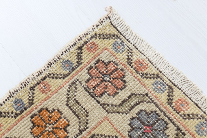 May include: Close-up of a corner of a beige rug with a floral pattern. The rug has a fringe border and features a repeating design of orange, pink, and blue flowers.