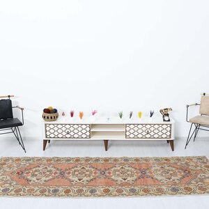 May include: A white entertainment center with two drawers and an open shelf sits on a beige rug with a floral pattern. Two black metal chairs with wooden armrests are placed on either side of the entertainment center. The entertainment center has a decorative lattice design on the front.