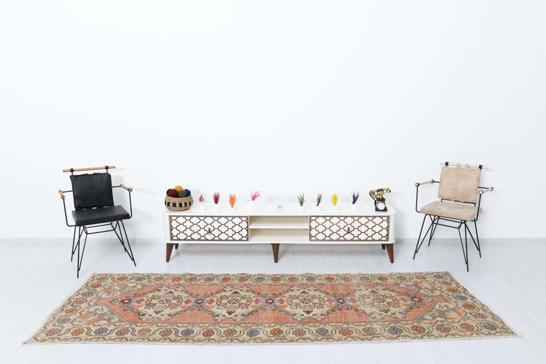 May include: A white entertainment center with two drawers and an open shelf sits on a beige rug with a floral pattern. Two black metal chairs with wooden armrests are placed on either side of the entertainment center. The entertainment center has a decorative lattice design on the front.