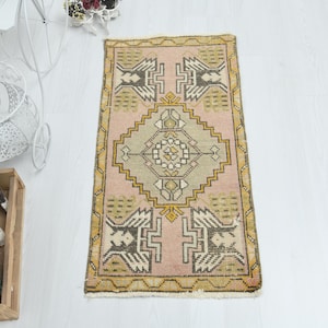May include: A rectangular area rug featuring a soft pink background with a central geometric diamond medallion in muted green and cream. The border and corners are decorated with stylized dark brown and yellow angular motifs and patterns.