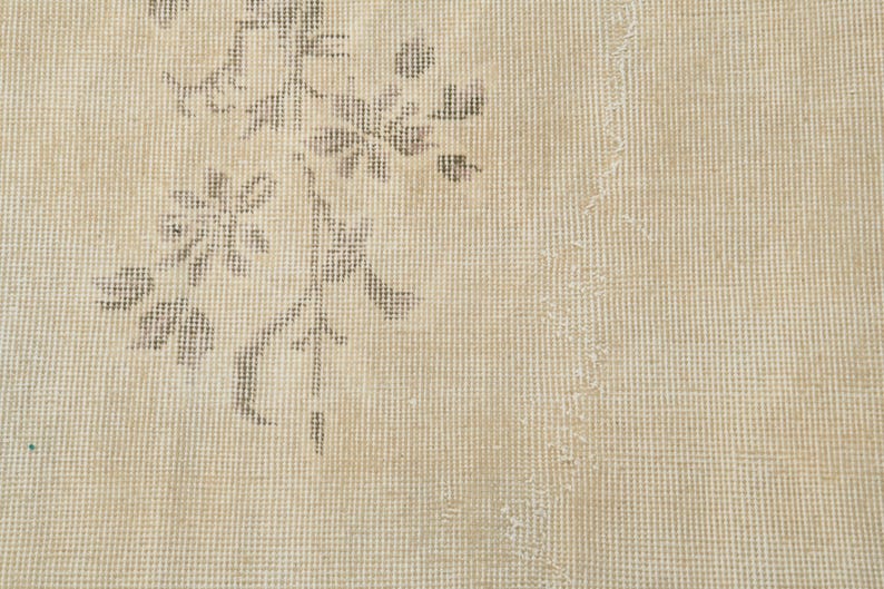 May include: Close-up of a beige rug with a floral pattern in shades of gray. The rug has a distressed, vintage look with visible wear and tear, adding to its character. The design is centered on the left side.