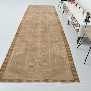 May include: A rectangular, beige-colored runner rug with a detailed, geometric pattern. The rug has a darker brown border and is placed on a white surface. The rug's design features floral and geometric motifs.