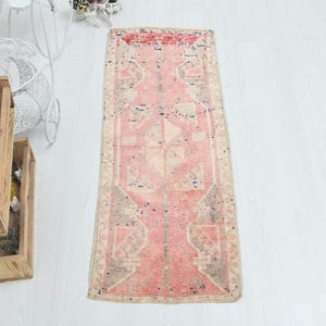May include: A rectangular runner rug featuring a faded pink central field with geometric patterns in cream, grey, and dark blue. The rug has a distressed texture and a decorative border with intricate motifs, showcasing a worn, vintage appearance.