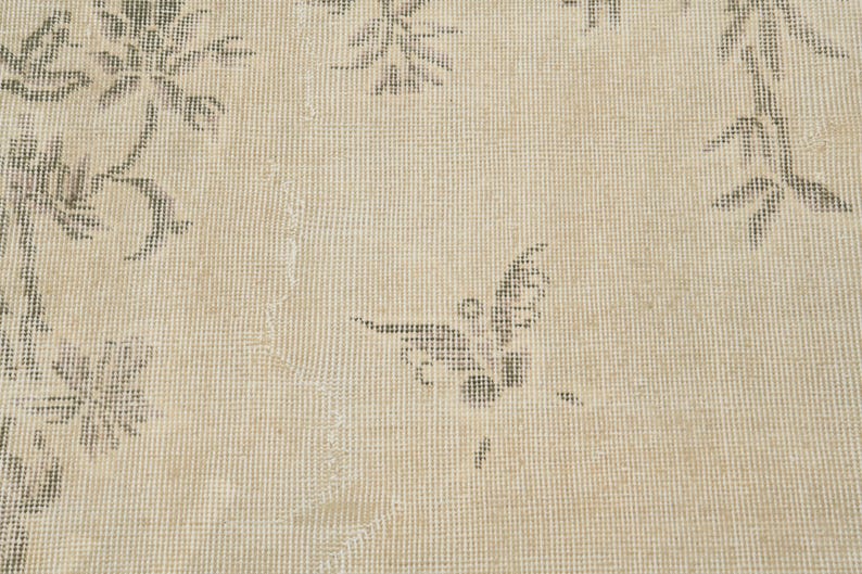 May include: Close-up of a beige rug with a floral pattern and a butterfly design. The rug has a distressed look with faded colors and a textured surface. The floral and butterfly motifs are in shades of gray.