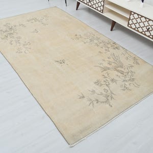May include: Beige rectangular rug with a floral and bird design in shades of gray. The rug has a cream-colored background and a border. The design features a bird perched on a branch with leaves and flowers.