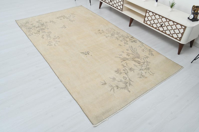 May include: Beige rectangular rug with a floral and bird design in shades of gray. The rug has a cream-colored background and a border. The design features a bird perched on a branch with leaves and flowers.