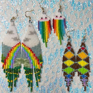 May include: A collection of beaded earrings in various designs. One pair features a cloud and rainbow design, another has a rainbow with a smiling face, and the last pair has a geometric pattern in brown, yellow, green, and blue. All earrings have gold-colored hooks.