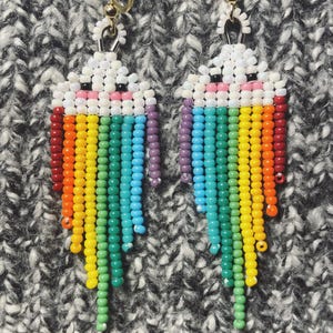 Puede incluir: Un par de pendientes de cuentas con forma de nube con un arcoíris. La parte superior de los pendientes es blanca con detalles negros y rosas. El arcoíris incluye rojo, naranja, amarillo, verde, azul y morado.