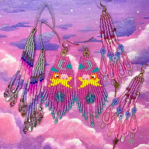Könnte beinhalten: Eine Auswahl an Ohrringen aus Perlen in verschiedenen Designs. Die Ohrringe bestehen aus rosa, lila und blauen Perlen, wobei einige Designs Sterne und Monde enthalten. Der Hintergrund ist ein bemalter Himmel mit Wolken.