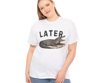 Könnte beinhalten: Weißes T-Shirt mit dem Wort "LATER" über einer Illustration eines Alligators. Der Alligator ist in Grau- und Brauntönen gehalten und liegt auf einem Fleck aus beigem Boden. Das T-Shirt wird von einer Person getragen.