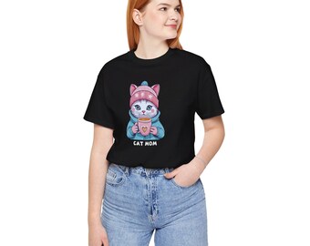 May include: Black t-shirt featuring a cute cat illustration wearing a pink hat and holding a coffee cup with a heart. The text "CAT MOM" is printed below the cat. The shirt is made of a soft material.