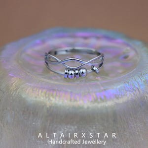May include: A silver ring with an intricate design. The ring has interwoven bands and small, spherical beads. The ring is presented on a pearlescent surface. The text "ALTAIRXSTAR Handcrafted Jewellery" is at the bottom of the image.