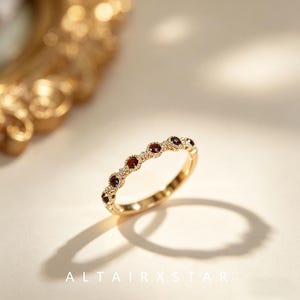 May include: A gold band ring featuring a series of small, round, deep red garnet stones set within individual circular frames, accented by tiny clear stones between each setting. The metal has a polished finish and the text ALTAIRXSTAR is visible.