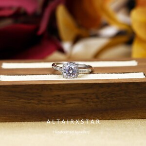 May include: A silver ring with a large, round, clear gemstone at the center, surrounded by smaller stones. The ring is set on a wooden jewelry box. The text "ALTAIRXSTAR Handcrafted Jewellery" is visible.