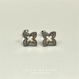 May include: A pair of silver stud earrings. Each earring has a four-petal flower design with a filigree pattern and a white centre. The earrings are from ALTAIRXSTAR, Handcrafted Jeweller.