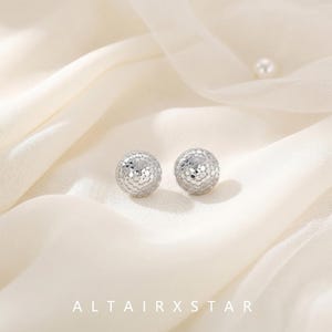 May include: A pair of silver-toned spherical stud earrings featuring a textured, faceted surface that reflects light. The earrings are positioned on a soft, draped off-white fabric. The text ALTAIRXSTAR is printed in white at the bottom.