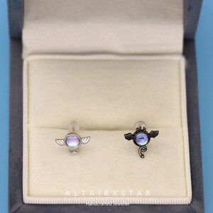 May include: A pair of stud earrings with a celestial design. One earring is silver-toned, the other is black. Each features a central iridescent stone, surrounded by small wings. The earrings are displayed in a gray jewelry box. The text "ALTAIRXSTAR Handcrafted Jeweller" is at the bottom.