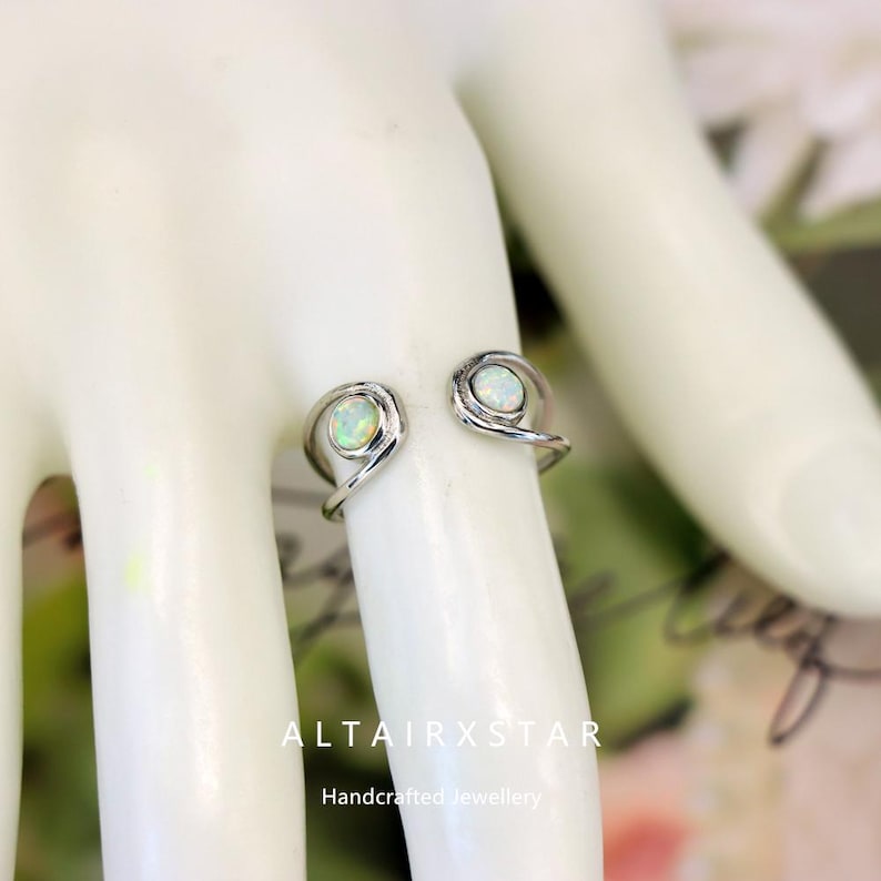 May include: A silver-colored open ring with two opal gemstones. The ring has a unique design with swirling details around the gemstones. The ring is handcrafted. The text "ALTAIRXSTAR" and "Handcrafted Jewellery" are visible.