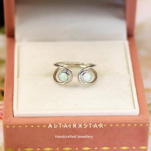 May include: A silver ring with two opal stones, set in a unique open design. The ring is displayed in a pink jewelry box with the text "ALTAIRXSTAR" and "Handcrafted Jewellery". The opals have a subtle, iridescent glow.
