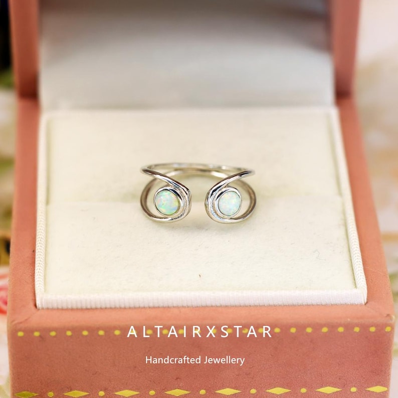 May include: A silver ring with two opal stones, set in a unique open design. The ring is displayed in a pink jewelry box with the text "ALTAIRXSTAR" and "Handcrafted Jewellery". The opals have a subtle, iridescent glow.