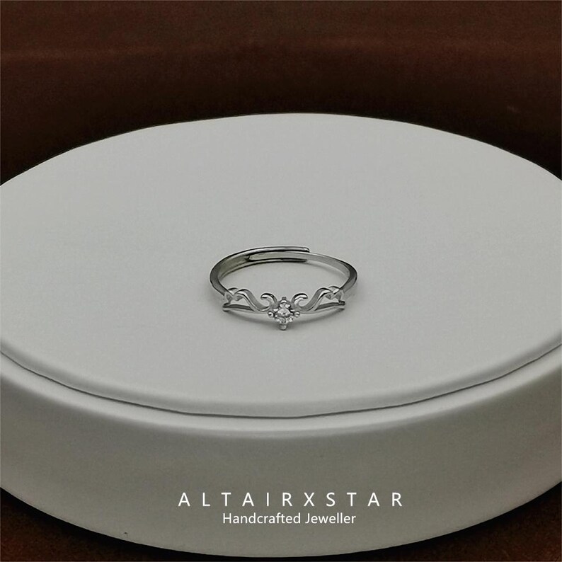 May include: A silver-toned adjustable ring with a decorative design and a central clear gemstone. The ring features a stylized wave pattern on either side of the gemstone. The text "ALTAIRXSTAR Handcrafted Jeweller" is at the bottom.