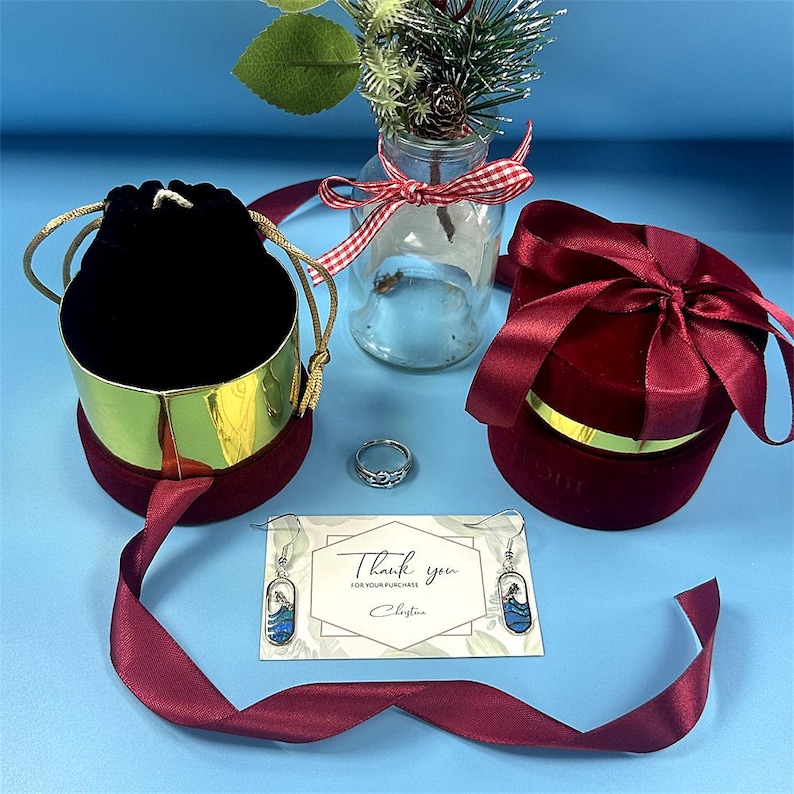 May include: An assortment of jewelry and gift packaging on a blue surface. Includes a silver ring, earrings with blue accents, a black velvet bag, and a red velvet gift box. A card reads "Thank you for your purchase".