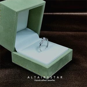 May include: A silver ring with a small square gemstone is displayed inside an open, green velvet jewelry box. The ring has a unique design with a curved band and a decorative element. The box is lined with white satin. The text "ALTAIRXSTAR Handcrafted Jeweller" is at the bottom.