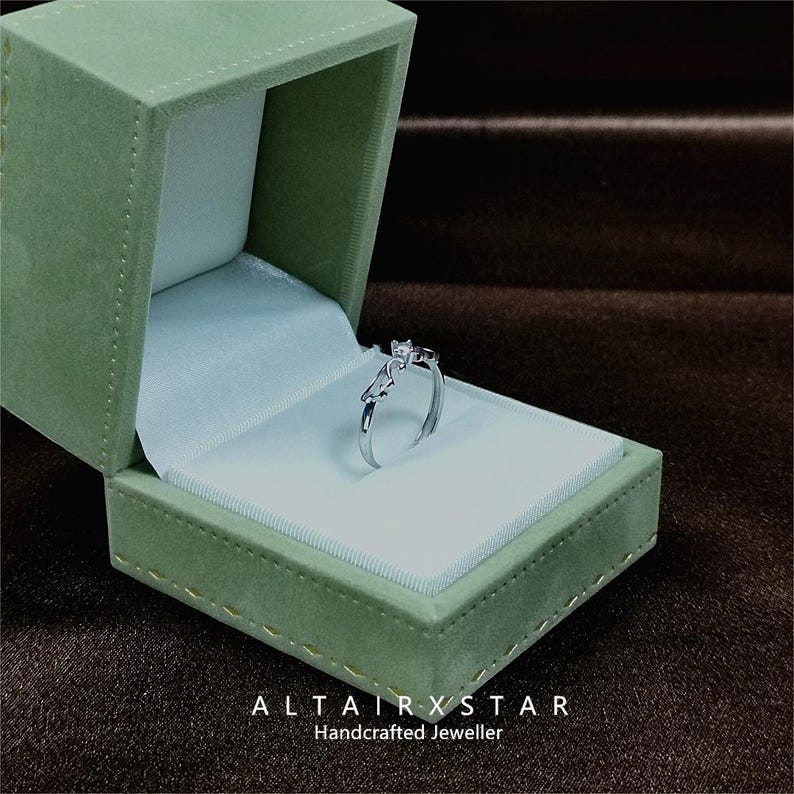 May include: A silver ring with a small square gemstone is displayed inside an open, green velvet jewelry box. The ring has a unique design with a curved band and a decorative element. The box is lined with white satin. The text "ALTAIRXSTAR Handcrafted Jeweller" is at the bottom.
