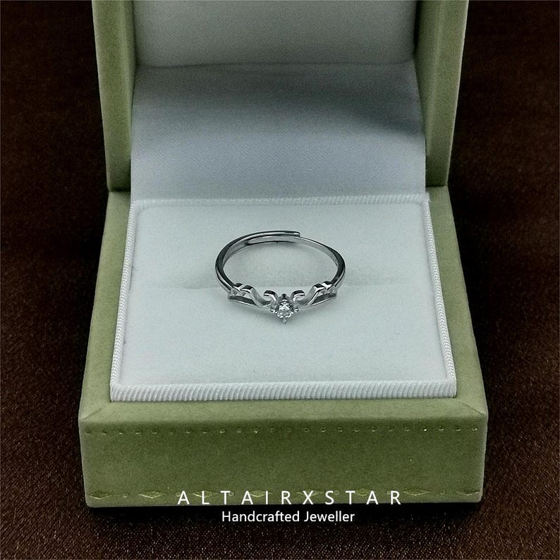 May include: A silver ring with a small, clear gemstone at the center, set in an ornate design. The ring is displayed inside a green jewelry box with a white interior. The text "ALTAIRXSTAR Handcrafted Jeweller" is at the bottom.