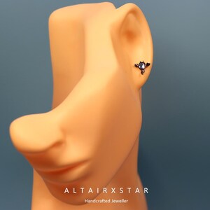 May include: A close-up of a small, dark-colored earring with a circular, light-colored stone. The earring is on a mannequin's ear. The text "ALTAIRXSTAR Handcrafted Jeweller" is at the bottom of the image.
