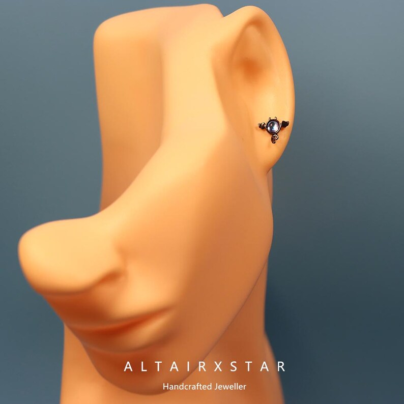 May include: A close-up of a small, dark-colored earring with a circular, light-colored stone. The earring is on a mannequin's ear. The text "ALTAIRXSTAR Handcrafted Jeweller" is at the bottom of the image.