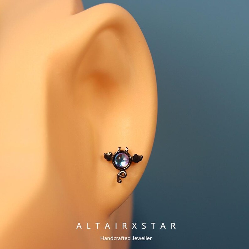 May include: A black stud earring with a devil design. The earring features a round, iridescent stone at the center, with small horns, wings, and a tail. The text "ALTAIRXSTAR Handcrafted Jeweller" is at the bottom.