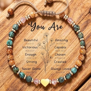 Könnte beinhalten: Ein Armband mit türkisfarbenen, braunen und rosa Perlen. Das Armband hat einen gelben Herzanhänger und liegt auf einer Holzoberfläche mit den Worten "You Are" und inspirierenden Worten wie "Beautiful" und "Amazing".