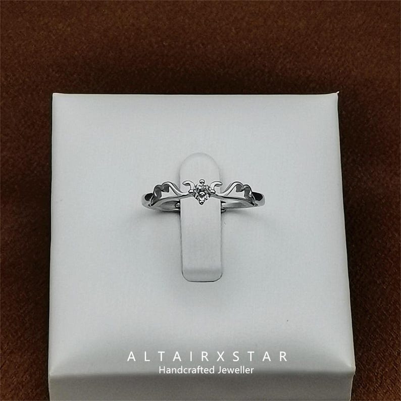 May include: A silver ring with a small, clear gemstone at the center. The ring has a decorative design on either side of the gemstone. The ring is displayed on a white stand, with the text "ALTAIRXSTAR Handcrafted Jeweller" at the bottom.